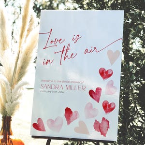 May include: A white sign with the text "Love is in the air" in red script. The sign is decorated with pink and red watercolor hearts. The text "Welcome to the Bridal Shower of SANDRA MILLER February 14th 20xx" is printed below the hearts.