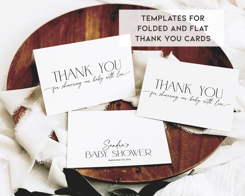 May include: Three white thank you cards with black calligraphy text. The text reads "Thank you for showering our baby with love". The cards are on a wooden surface with a white fabric overlay.