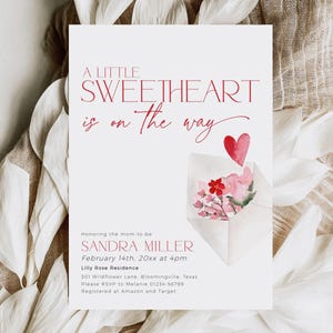 May include: A pink and white baby shower invitation with a red heart and flowers in an envelope. The text reads "A Little Sweetheart is on the way" and includes the name Sandra Miller, the date February 14th, and the time 4pm. The invitation also includes the location Lilly Rose Residence, the address 301 Wildflower Lane, Bloomingville, Texas, and the RSVP information.