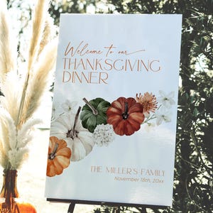May include: A Thanksgiving dinner welcome sign with the text "Welcome to our Thanksgiving Dinner" in copper-colored script. The sign features illustrations of pumpkins and flowers in shades of orange, green, and white.