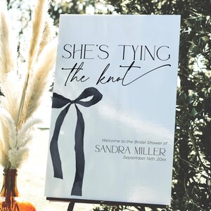 Op de afbeelding: Een wit bord met zwarte tekst die "SHE'S TYING the knot" luidt met een zwarte strik. Onder de tekst staat "Welcome to the Bridal Shower of SANDRA MILLER September 14th 20xx".