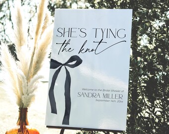 Puede incluir: Un cartel blanco con texto negro que dice "SHE'S TYING the knot" con un lazo negro. Debajo del texto se lee "Welcome to the Bridal Shower of SANDRA MILLER September 14th 20xx".