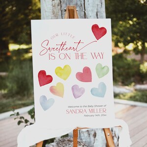 May include: A white sign with the text "Our Little Sweetheart Is On The Way" in red script. The sign is decorated with colorful watercolor hearts and the text "Welcome to the Baby Shower of Sandra Miller February 14th 20xx".
