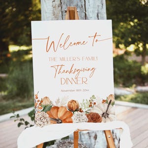 May include: A white sign on an easel with the words "Welcome to The Miller's Family Thanksgiving Dinner" in elegant script. The sign is decorated with fall-themed floral and pumpkin illustrations. The date "November 18th, 20xx" is also displayed.