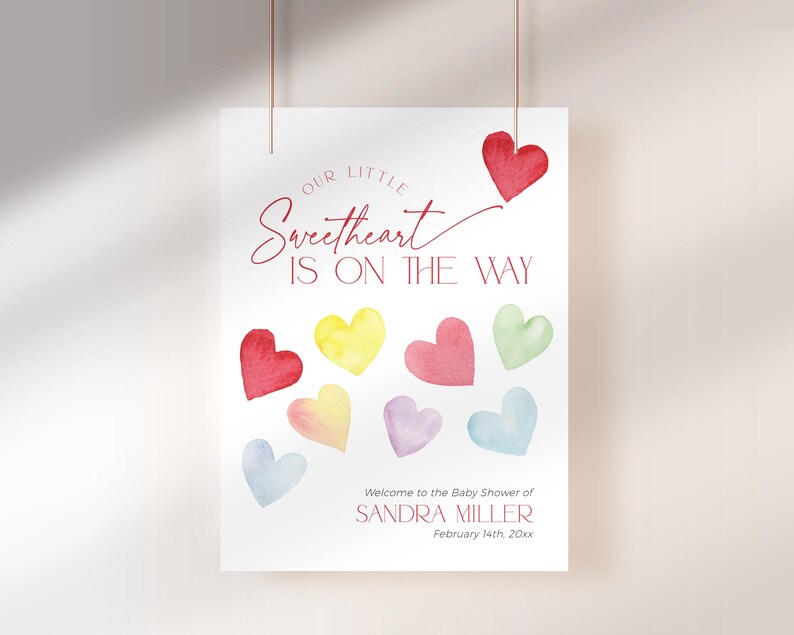 May include: A white card with the text "Our Little Sweetheart Is On The Way" in red script. The card is decorated with colorful watercolor hearts. The text "Welcome to the Baby Shower of Sandra Miller February 14th, 20xx" is printed at the bottom of the card.