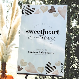 K&ouml;nnte beinhalten: Ein wei&szlig;es Schild mit schwarzem Text, das eine Babyparty ank&uuml;ndigt. Der Text lautet "Unser kleiner Liebling ist unterwegs". Das Schild enth&auml;lt auch den Namen "Sandras Babyparty" und das Datum "14. Februar 20xx". Das Schild ist mit Herzen in verschiedenen Braunt&ouml;nen und Schwarz verziert.