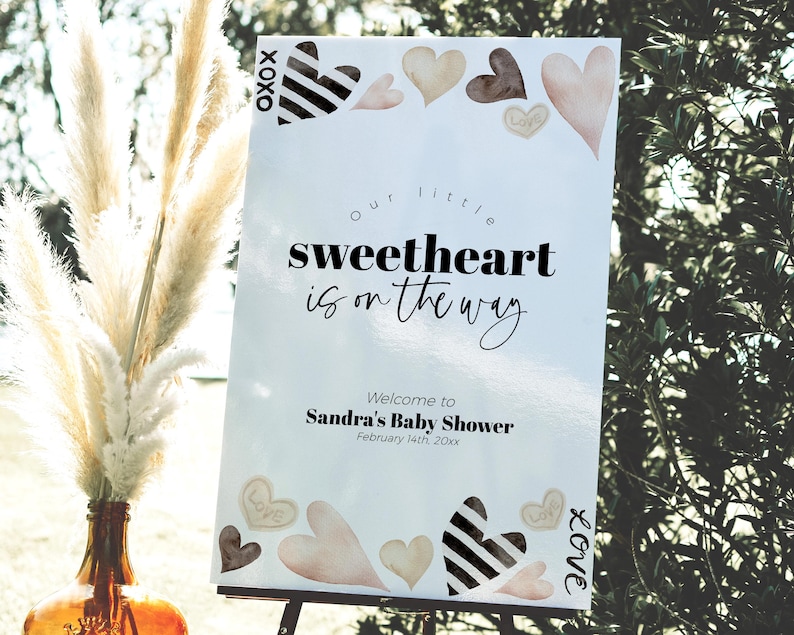 K&ouml;nnte beinhalten: Ein wei&szlig;es Schild mit schwarzem Text, das eine Babyparty ank&uuml;ndigt. Der Text lautet "Unser kleiner Liebling ist unterwegs". Das Schild enth&auml;lt auch den Namen "Sandras Babyparty" und das Datum "14. Februar 20xx". Das Schild ist mit Herzen in verschiedenen Braunt&ouml;nen und Schwarz verziert.