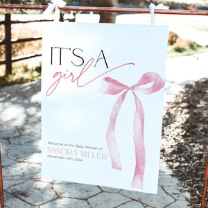 May include: It's a girl baby shower sign featuring a pink watercolour bow and the text "Welcome to the Baby Shower of Sandra Miller, December 14th 20xx".