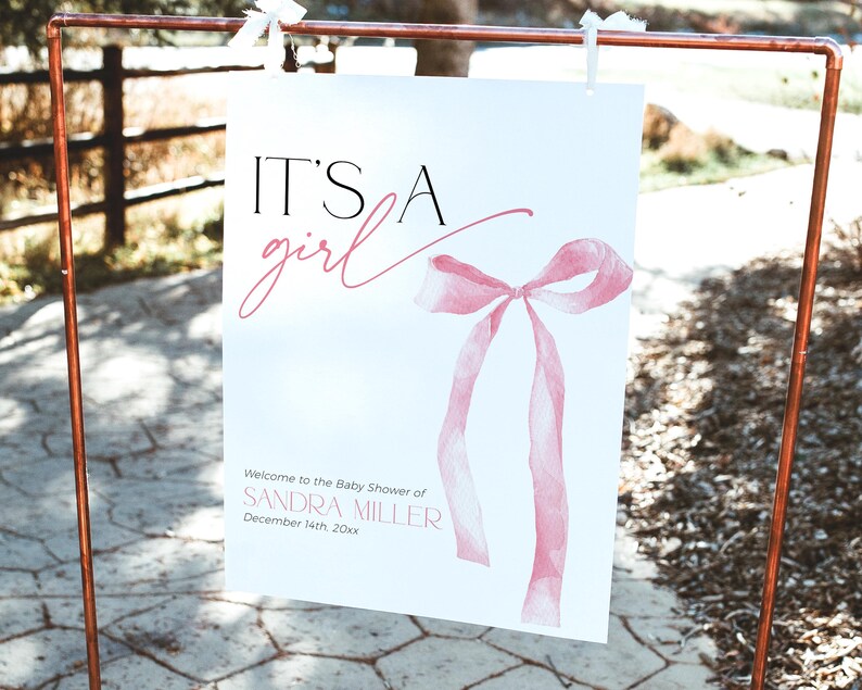 May include: It's a girl baby shower sign featuring a pink watercolour bow and the text "Welcome to the Baby Shower of Sandra Miller, December 14th 20xx".