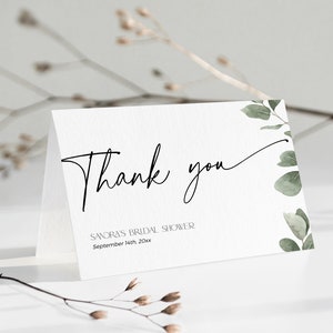 Może przedstawiać: Biała kartka z podziękowaniem z czarną kaligrafią, na kt&oacute;rej widnieje napis "Dziękuję" oraz tekst "Sandra's Bridal Shower September 14th 20xx". Kartka ma zielony akwarelowy wz&oacute;r kwiatowy po prawej stronie.
