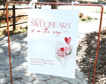 Könnte beinhalten: Ein weißes Schild mit rotem Text, das eine Babyparty für Sandra Miller ankündigt. Das Schild sagt "A Little Sweetheart is on the way" und enthält eine Aquarell-Illustration eines rosa Umschlags mit Blumen und einem roten Herzen.