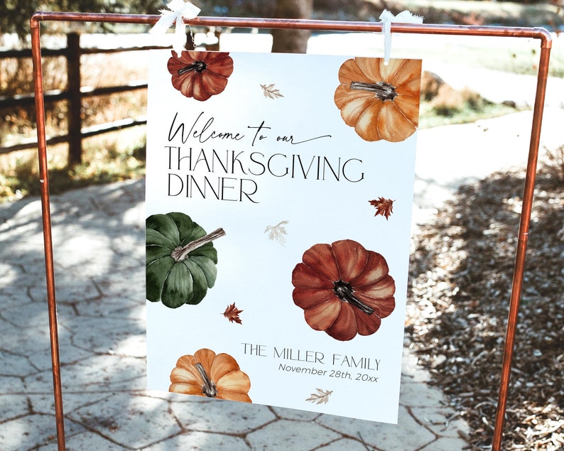May include: Thanksgiving dinner sign with watercolor pumpkins in shades of orange, green, and brown. The sign reads "Welcome to our Thanksgiving Dinner" and includes the family name and date. The sign is displayed on a copper-colored metal stand.