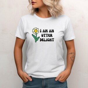 May include: White t-shirt with a smiling flower graphic and the text "I AM AN UTTER DELIGHT" in black. The flower has a yellow center, white petals, and a green stem and leaves. The shirt is short-sleeved.