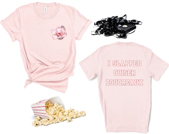 May include: Two pink t-shirts, one with a floral design and the other with the text "I SLAPPED OUISER BOUDREAUX" in red. A bag of popcorn is spilled on a white surface.