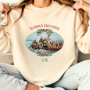 May include: A cream-colored sweatshirt with a vintage-style illustration of a boat full of people wearing Santa hats. The text "Season's Greetings 1776" is printed on the sweatshirt.