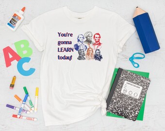 May include: White t-shirt with a blue and red graphic that says "You're gonna LEARN today!" and features images of historical figures. The shirt is surrounded by school supplies, including a pencil, scissors, crayons, markers, and a composition book.