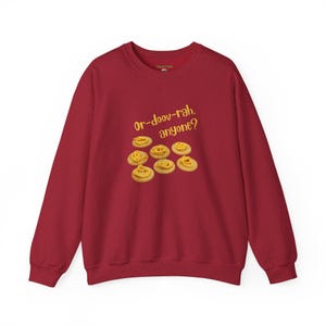 Könnte beinhalten: Roter Pullover mit dem Text "Or-doov-rah. anyone?" und einer Grafik von acht kleinen Keksen mit gelber Glasur.