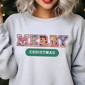 May include: A light blue sweatshirt with the words "MERRY CHRISTMAS" in a colorful, collegiate-style design. The word "MERRY" is in a red and white font, and the word "CHRISTMAS" is in a green oval.
