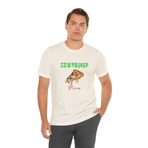 Puede incluir: Una camiseta blanca con un gráfico verde de "Cowabunga Pizza" que presenta una rebanada de pizza con pepperoni rojo, pimientos verdes y queso derretido.