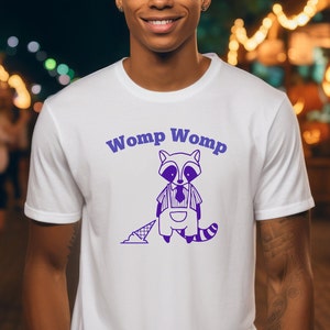 Puede incluir: Camiseta blanca con un gráfico de mapache morado y el texto "Womp Womp" impreso en la parte delantera.