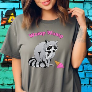 Può includere: Una maglietta grigia con il testo "Womp Womp" in lettere rosa. Un procione dei cartoni animati è raffigurato mentre guarda tristemente accanto a un cono gelato caduto.