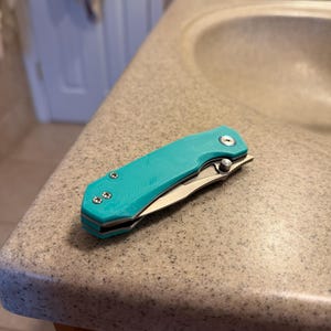 May include: A teal folding pocket knife with a silver blade and three silver screws. The knife is open, revealing the blade. The handle has a textured surface. The knife is resting on a speckled countertop.