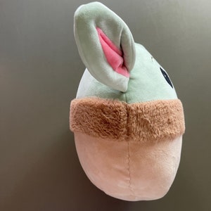 May include: A plush toy shaped like a character from the Star Wars franchise. The toy is green with a brown fur trim and has large pink ears.