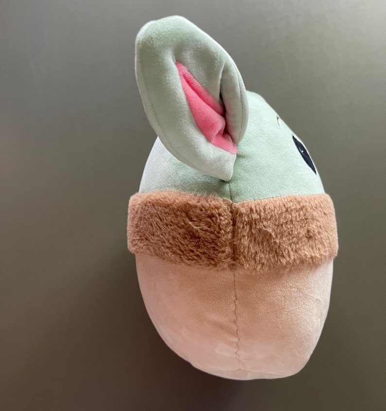 May include: A plush toy shaped like a character from the Star Wars franchise. The toy is green with a brown fur trim and has large pink ears.