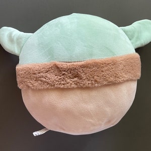 May include: A plush toy shaped like a character with large ears and a brown fur trim. The toy is light green and has a light brown bottom.