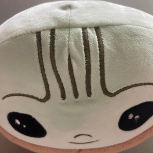 May include: A plush toy shaped like a green alien with a smiling face and black eyes. The toy has a brown stitched line across the forehead.