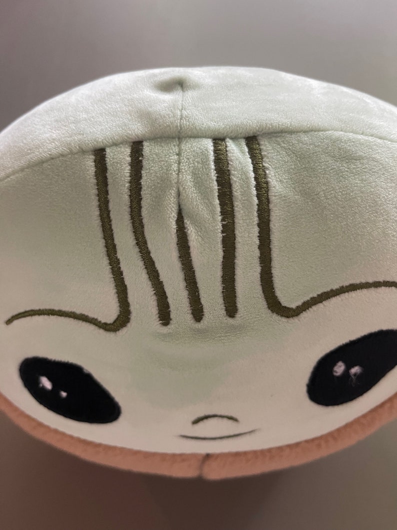 May include: A plush toy shaped like a green alien with a smiling face and black eyes. The toy has a brown stitched line across the forehead.