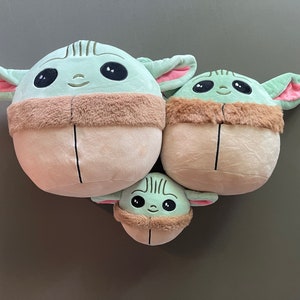May include: Three plush toys shaped like the character Baby Yoda from the Star Wars series. The toys are green and brown with large eyes and a small mouth. The largest toy is about 12 inches tall, the middle toy is about 8 inches tall, and the smallest toy is about 5 inches tall.