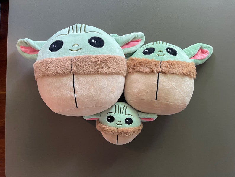 May include: Three plush toys shaped like the character Baby Yoda from the Star Wars series. The toys are green and brown with large eyes and a small mouth. The largest toy is about 12 inches tall, the middle toy is about 8 inches tall, and the smallest toy is about 5 inches tall.