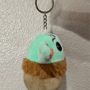 May include: A green and brown plush keychain with a cartoon face and a silver chain. The keychain is shaped like a small creature with a furry brown collar.