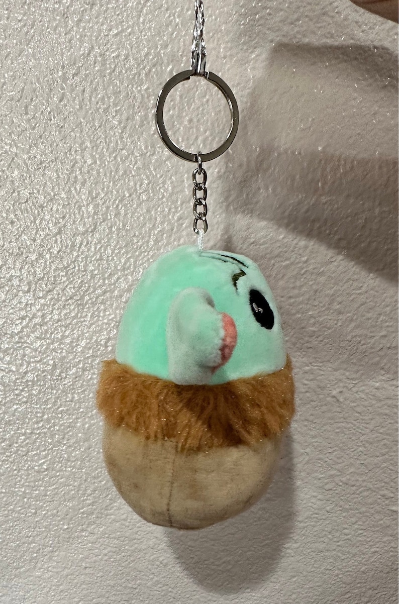 May include: A green and brown plush keychain with a cartoon face and a silver chain. The keychain is shaped like a small creature with a furry brown collar.