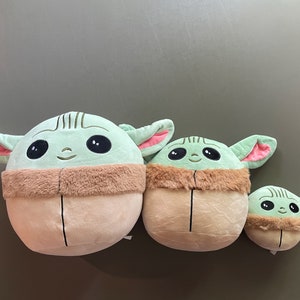 May include: Three plush toys shaped like the character Baby Yoda from the Star Wars series. The toys are green with large eyes and brown fur around their necks. The toys are different sizes, with the largest being about 12 inches tall.