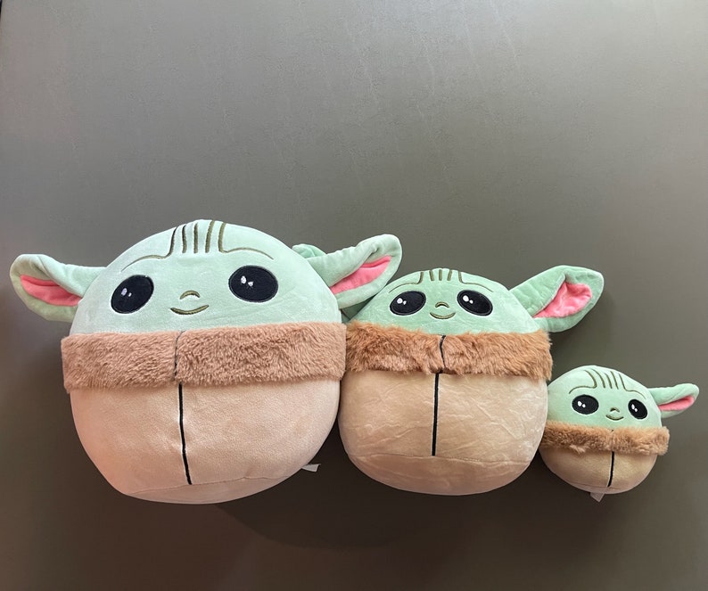 May include: Three plush toys shaped like the character Baby Yoda from the Star Wars series. The toys are green with large eyes and brown fur around their necks. The toys are different sizes, with the largest being about 12 inches tall.