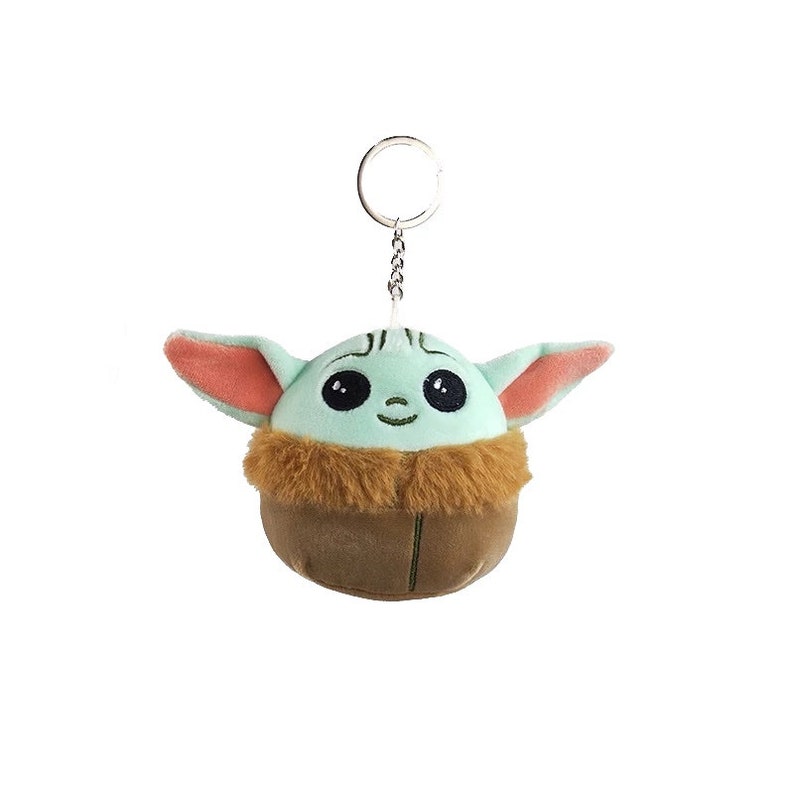 May include: A plush keychain shaped like a green and brown character with large eyes and a small smile. The character has large ears and a furry brown collar. The keychain is attached to a silver metal ring.