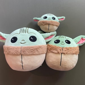 May include: Three plush toys shaped like the character Baby Yoda from the Star Wars series. The toys are green with large eyes and brown fur around their necks. The largest toy is about 10 inches tall, the middle toy is about 7 inches tall, and the smallest toy is about 5 inches tall.