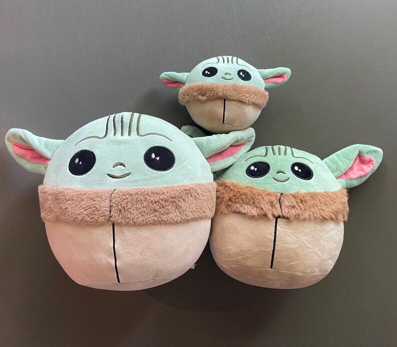 May include: Three plush toys shaped like the character Baby Yoda from the Star Wars series. The toys are green with large eyes and brown fur around their necks. The largest toy is about 10 inches tall, the middle toy is about 7 inches tall, and the smallest toy is about 5 inches tall.