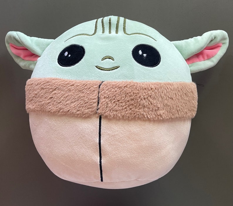 May include: A green plush toy shaped like a character from the Star Wars franchise. The toy has large eyes, a small mouth, and a brown fur collar.