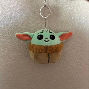May include: A green and brown plush keychain shaped like a character from the Star Wars franchise, with large eyes and a smiling face. The keychain is attached to a silver metal chain.