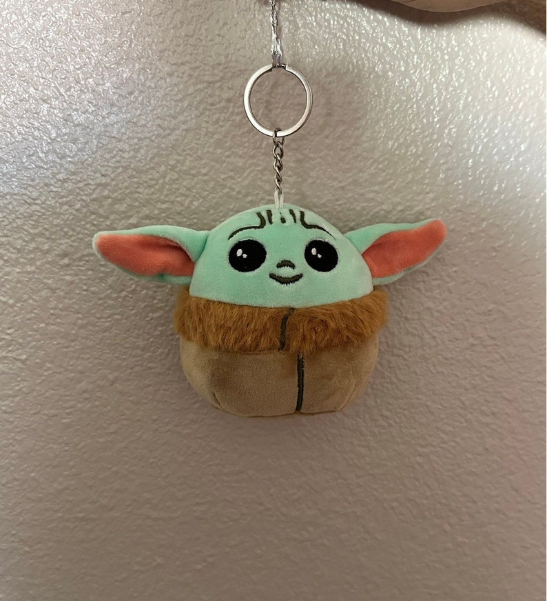 May include: A green and brown plush keychain shaped like a character from the Star Wars franchise, with large eyes and a smiling face. The keychain is attached to a silver metal chain.