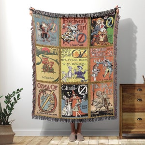 May include: A woven throw blanket featuring colorful illustrations of classic "Oz" book covers. The blanket displays various titles, including "Lost Princess of Oz" and "The Wonderful Wizard of Oz," with fringe detailing along the edges.