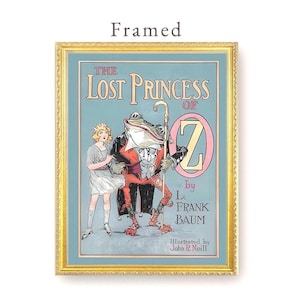 May include: Framed vintage illustration from "The Lost Princess of Oz" by L. Frank Baum. The artwork features a young girl and a frog-like character with a cane. The title and author's name are displayed in elegant typography, framed in a gold border.
