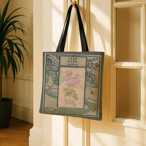 May include: A rectangular tote bag with black shoulder straps featuring a printed book cover design for Jane Eyre by Bront&euml;. The graphic displays a central pink rose illustration on a beige background, framed by dark green landscape borders.