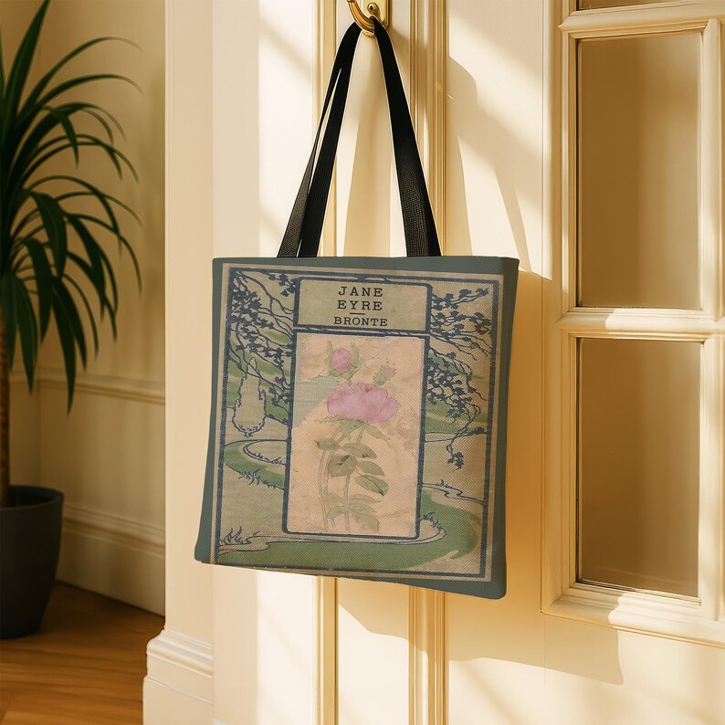 May include: A rectangular tote bag with black shoulder straps featuring a printed book cover design for Jane Eyre by Bront&euml;. The graphic displays a central pink rose illustration on a beige background, framed by dark green landscape borders.