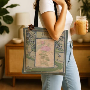 May include: A tote bag with black straps featuring a rectangular graphic print of a pink flower on a beige background, surrounded by a green landscape illustration and the text Jane Eyre Bronte in a dark blue border frame.