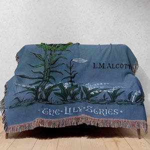 May include: A blue woven throw blanket with a floral design featuring lilies and the text "The Lily Series" and "L.M. Alcott".