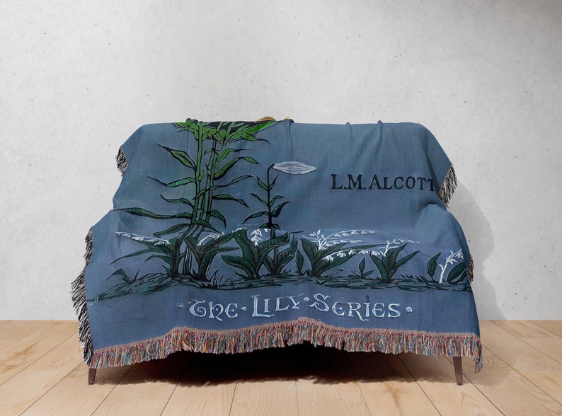May include: A blue woven throw blanket with a floral design featuring lilies and the text "The Lily Series" and "L.M. Alcott".